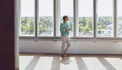 A woman stands alone in a spacious, empty room with expansive windows, creating a bright and airy atmosphere.