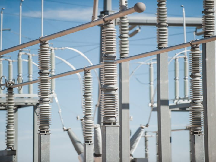 Close-up of an electrical substation with ceramic insulators, metal conductors, support structures, and power lines against a clear blue sky.