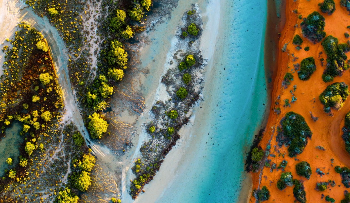 Aerial view of a coastal landscape with turquoise waterways, white sandy shores, green mangrove trees, and vibrant red-orange sand.