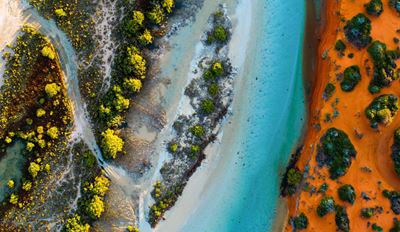Aerial view of a coastal landscape with turquoise waterways, white sandy shores, green mangrove trees, and vibrant red-orange sand.