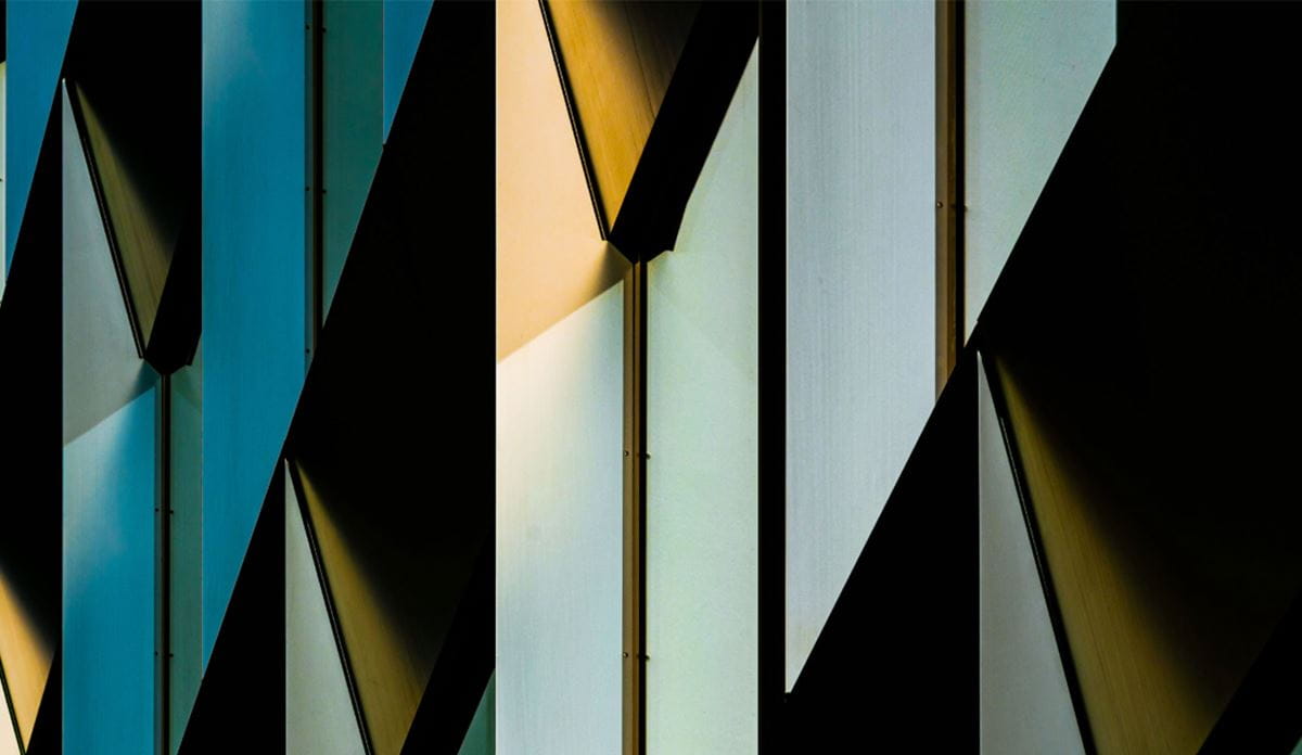 Abstract close-up of a modern building facade featuring geometric angular panels in teal, gold, and white tones with dramatic shadows.