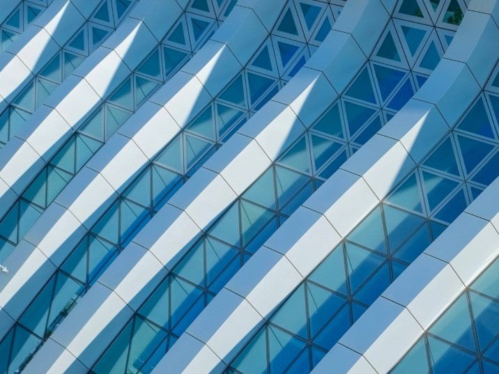 Close-up view of a curved modern building facade with diagonal white ribs and triangular blue glass panels, creating a repeating geometric pattern.