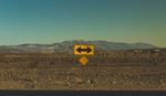 Yellow two-way traffic arrow sign in desert with mountains in background.