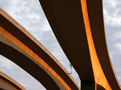 Upward view of curved concrete highway overpasses illuminated by warm golden sunlight, with layered elevated roadways sweeping across a cloudy sky.