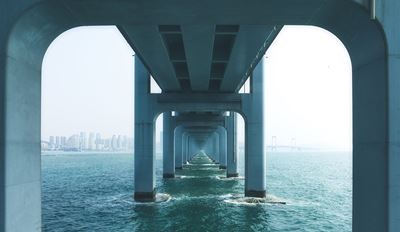 View from underneath a bridge showing concrete support pillars extending into blue-green water, with a city skyline visible in the distance.