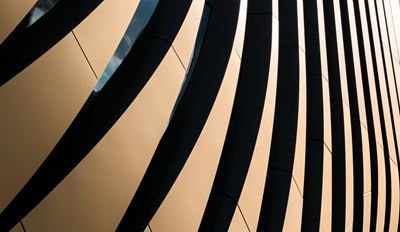 Close-up of a modern building facade featuring curved golden panels with bold dark shadow stripes, creating a dynamic abstract architectural pattern.