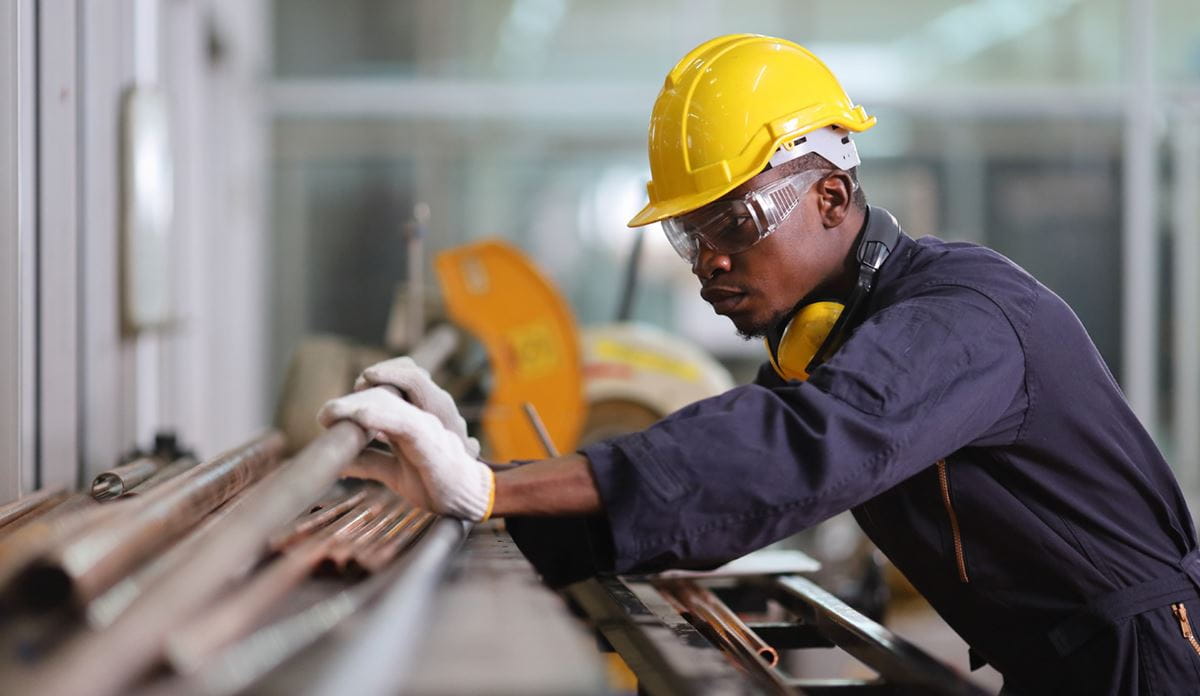 Worker wearing a yellow safety helmet, protective glasses, and gloves, inspecting metal pipes in an industrial workshop.