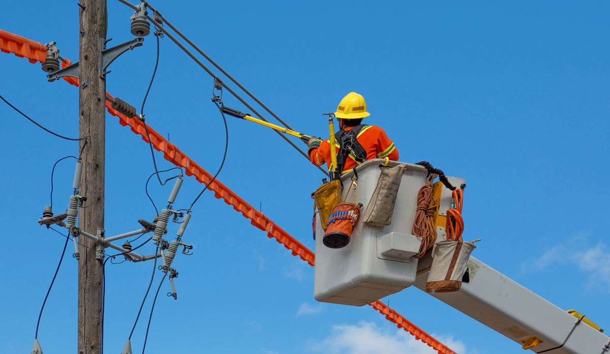 A utility worker in an orange safety uniform and helmet repairs power lines from a bucket lift.