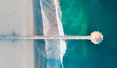 Aerial view of a sandy beach with a wooden pier extending into the water, surrounded by waves and beachgoers.