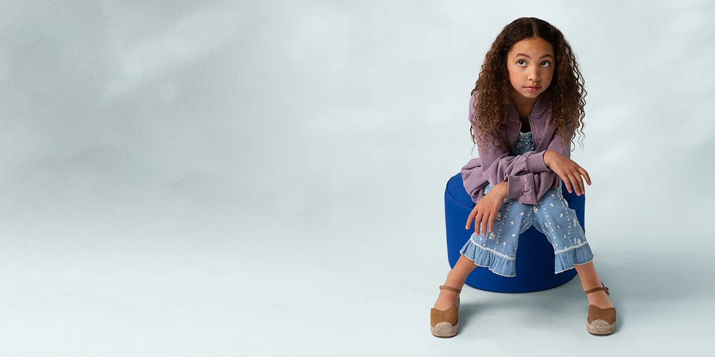 A young girl with curly hair sits thoughtfully on a blue ottoman against a light gray background, wearing a purple jacket, floral pants, and tan espadrilles.