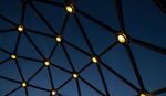 Low-angle view of a dark geometric dome framework illuminated by warm, circular lights against a deep blue night sky