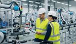 Two workers collaborate on an assembly line, surrounded by robotic arms assisting in the manufacturing process.