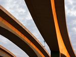 Upward view of curved concrete highway overpasses illuminated by warm golden sunlight, with layered elevated roadways sweeping across a cloudy sky.
