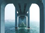 View from underneath a long concrete bridge extending over water toward a distant city skyline.