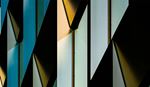 Abstract close-up of a modern building facade featuring geometric angular panels in teal, gold, and white tones with dramatic shadows.