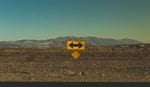 Yellow two-way traffic arrow sign in desert with mountains in background.