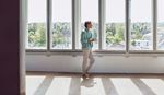 A woman stands alone in a spacious, empty room with expansive windows, creating a bright and airy atmosphere.