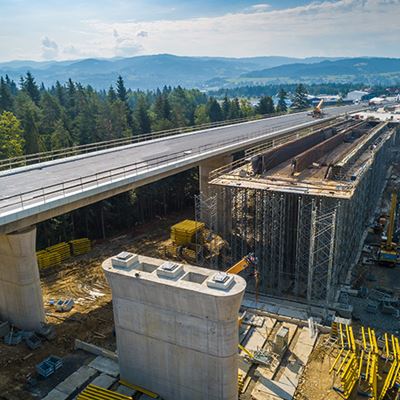 Aerial view of a highway bridge under construction over a forested area with mountains visible in the background.