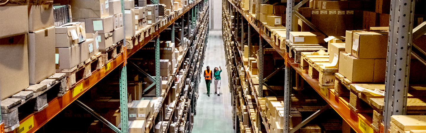 Large industrial warehouse with high metal storage racks filled with boxes and two people walking down the aisle