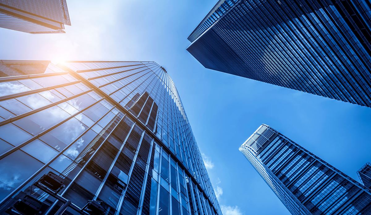 Looking up at modern glass skyscrapers against a clear blue sky, with sunlight reflecting off the buildings, conveying a sense of innovation and growth.