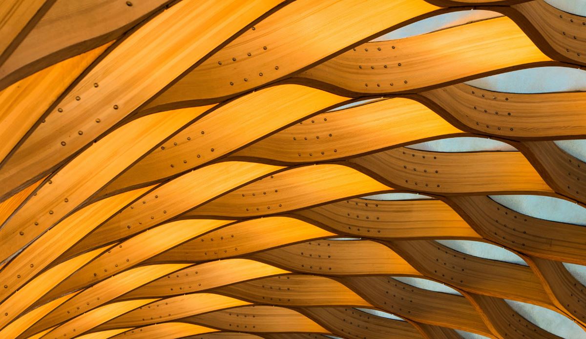 Close-up view of an abstract wooden ceiling structure with interlocking curved beams and visible bolts, illuminated with warm light.