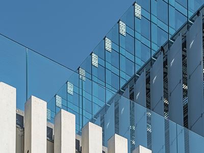 Low-angle view of a modern glass-paneled building facade reflecting blue sky, with white concrete columns in the foreground.