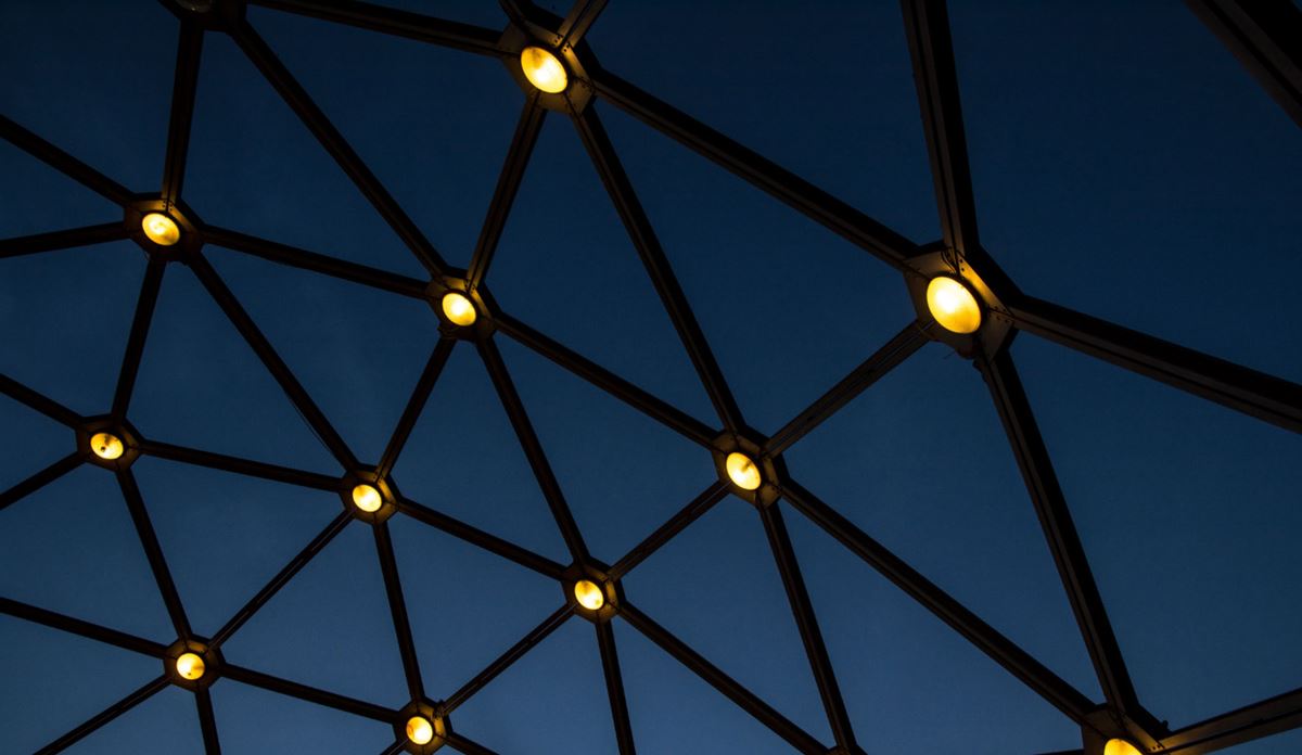 Low-angle view of a dark geometric dome framework illuminated by warm, circular lights against a deep blue night sky
