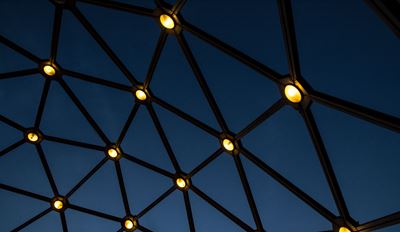 Low-angle view of a dark geometric dome framework illuminated by warm, circular lights against a deep blue night sky