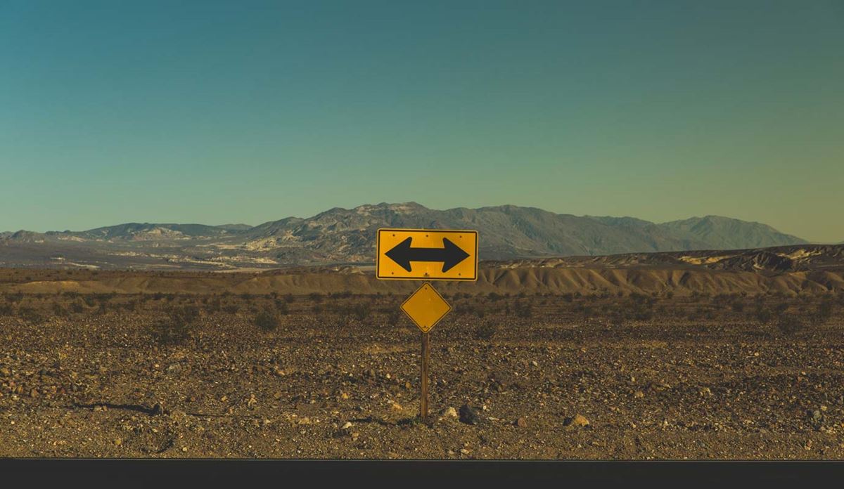 Yellow two-way traffic arrow sign in desert with mountains in background.