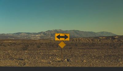 Yellow two-way traffic arrow sign in desert with mountains in background.