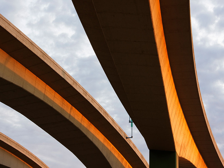 Upward view of curved concrete highway overpasses illuminated by warm golden sunlight, with layered elevated roadways sweeping across a cloudy sky.