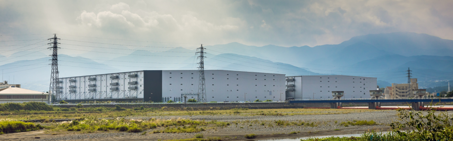 Large industrial warehouse complex with power transmission towers, set against a mountain backdrop near a riverbed in Odawara, Japan.