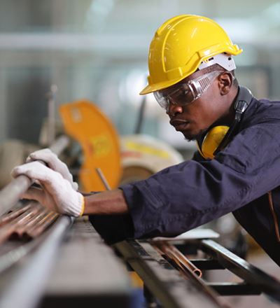 Worker wearing a yellow safety helmet, protective glasses, and gloves, inspecting metal pipes in an industrial workshop.