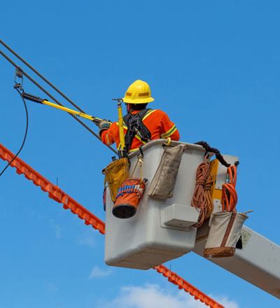 A utility worker in an orange safety uniform and helmet repairs power lines from a bucket lift.