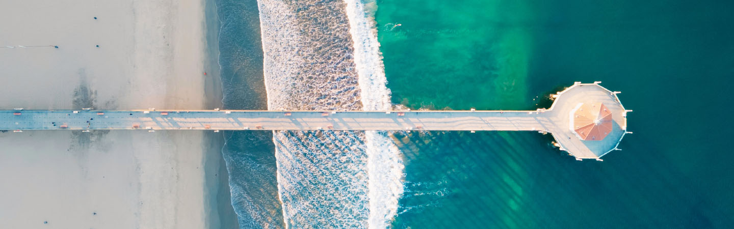 Aerial view of a sandy beach with a wooden pier extending into the water, surrounded by waves and beachgoers.