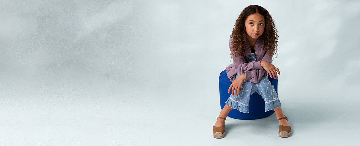 A young girl with curly hair sits thoughtfully on a blue ottoman against a light gray background, wearing a purple jacket, floral pants, and tan espadrilles.